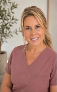 Smiling woman with blonde hair wearing pink scrubs, standing indoors with a potted plant in the background.
